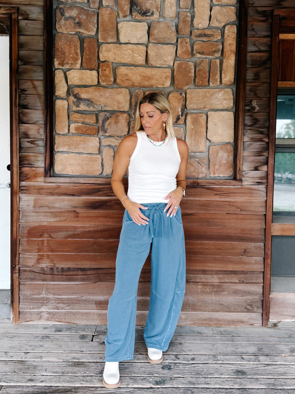 A woman stands on a rustic wooden porch in front of a stone wall, wearing a white sleeveless tank top, light blue wide-leg corduroy pants, and white sneakers.
