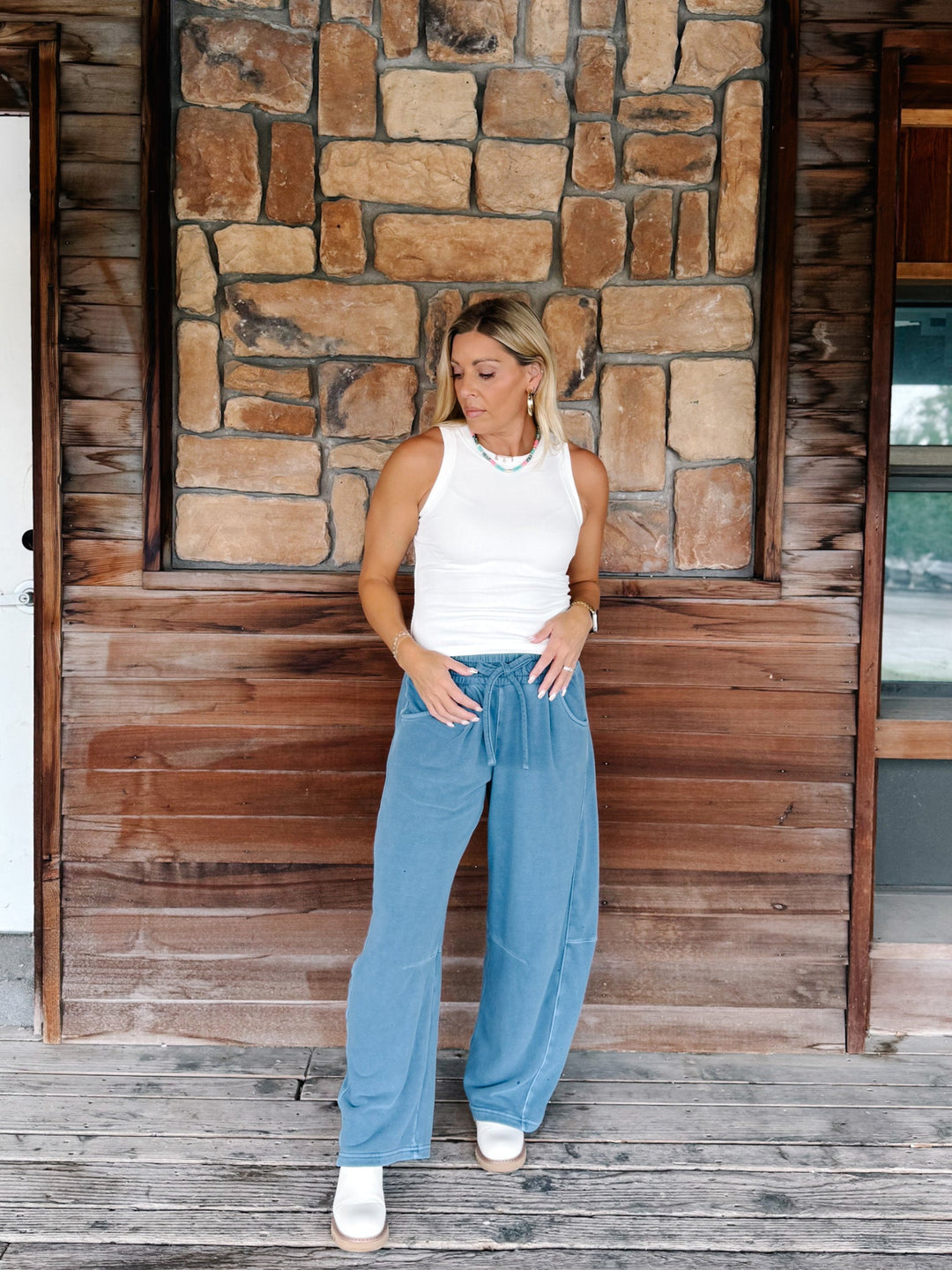 A woman stands on a rustic wooden porch in front of a stone wall, wearing a white sleeveless tank top, light blue wide-leg corduroy pants, and white sneakers.