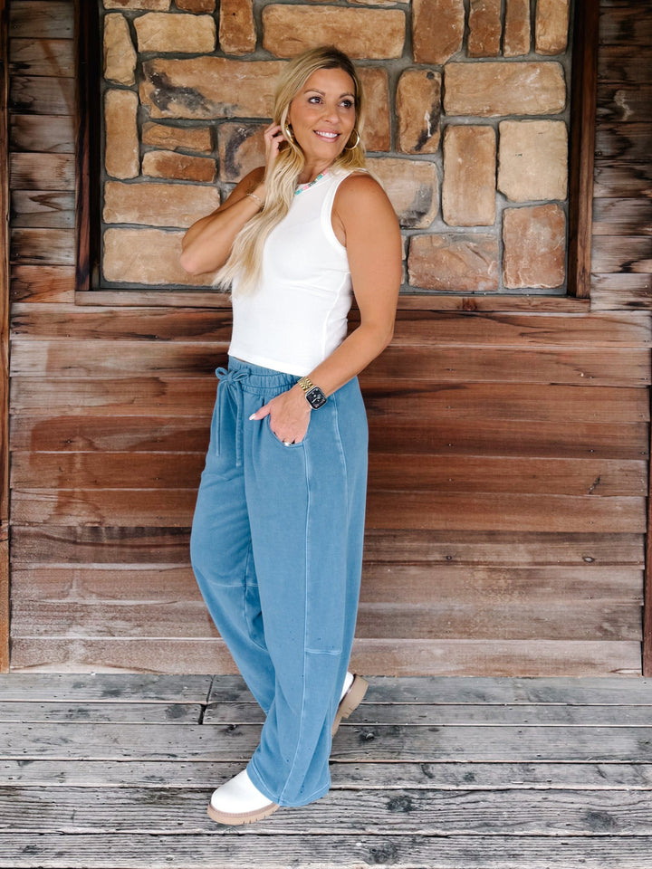A woman stands on a rustic wooden porch in front of a stone wall, wearing a white sleeveless top and light blue wide-leg drawstring pants with white sneakers.