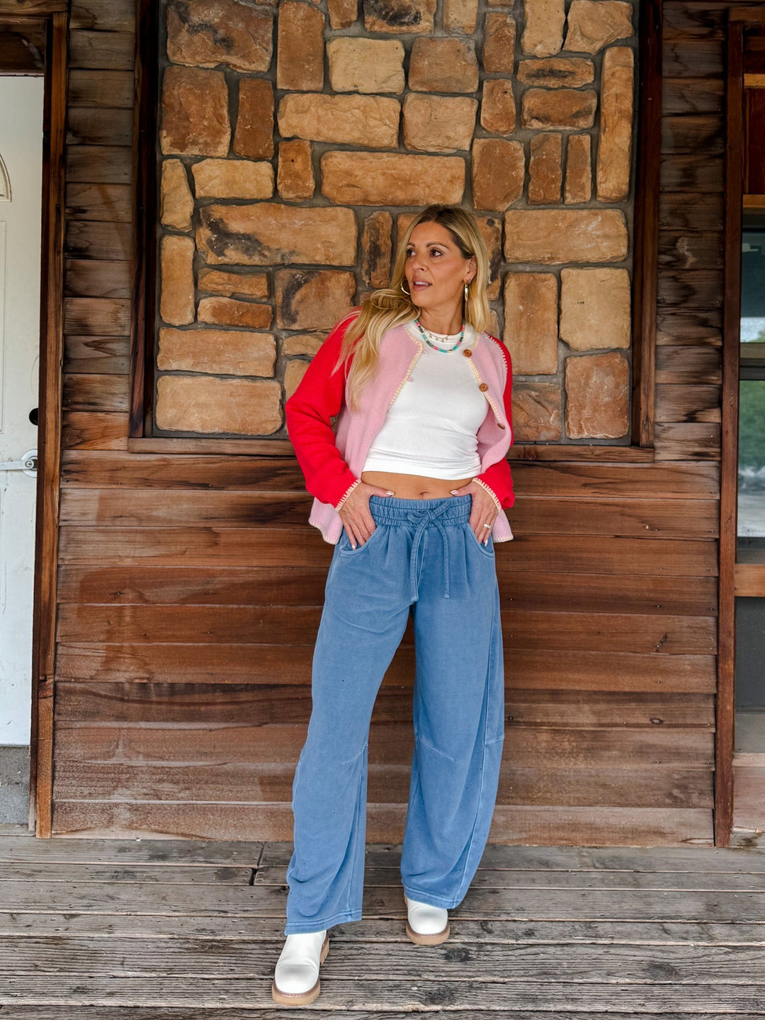 A woman stands on a wooden deck in front of a rustic stone and wood wall, wearing a bright pink and red cropped jacket over a white crop top, high-waisted blue wide-leg barrel pants, and white sneakers.