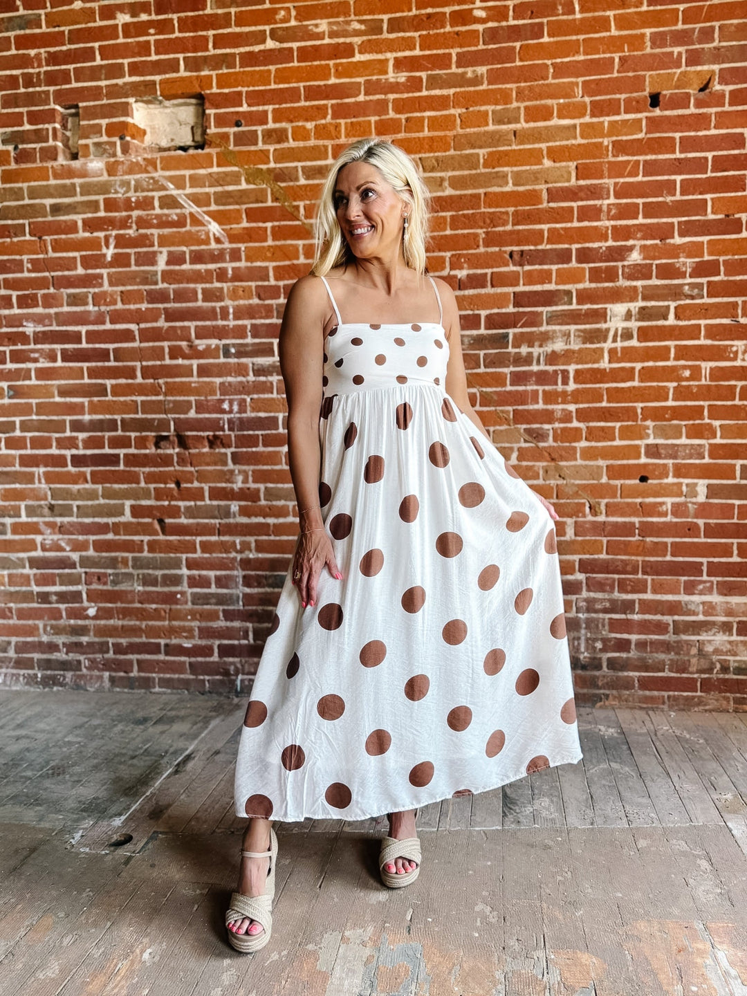 Woman wearing a white midi dress with oversized mocha polka dots, adjustable spaghetti straps, and an empire waist.