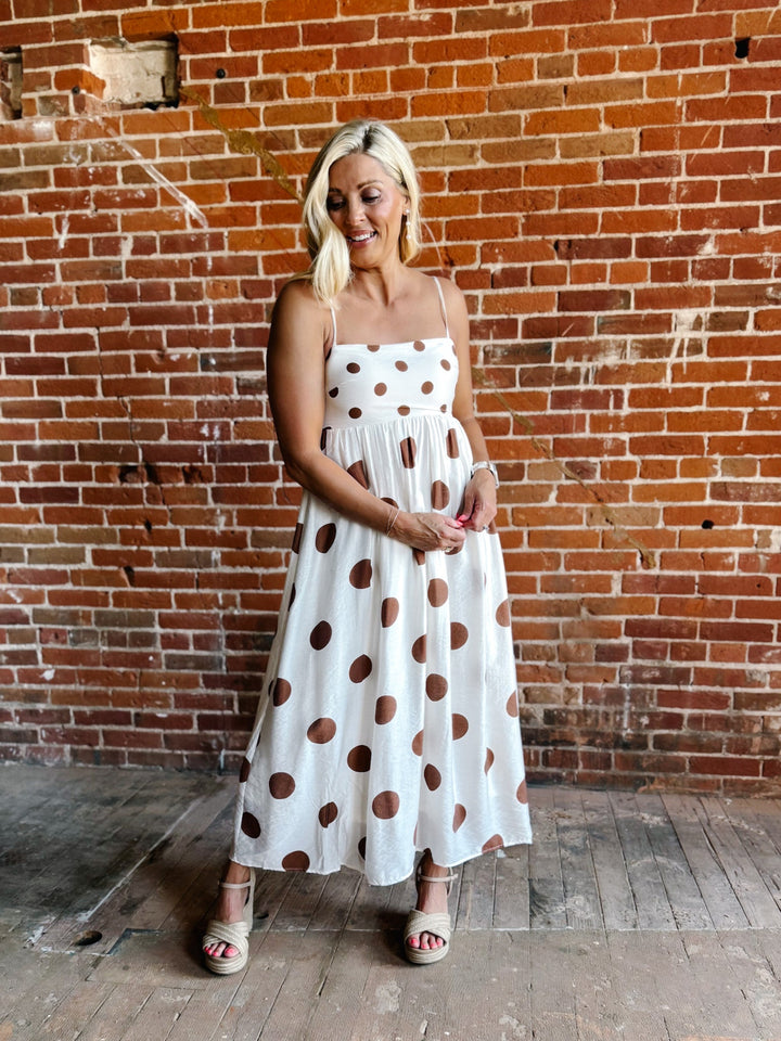 Woman wearing a white midi dress with oversized mocha polka dots, adjustable spaghetti straps, and an empire waist.