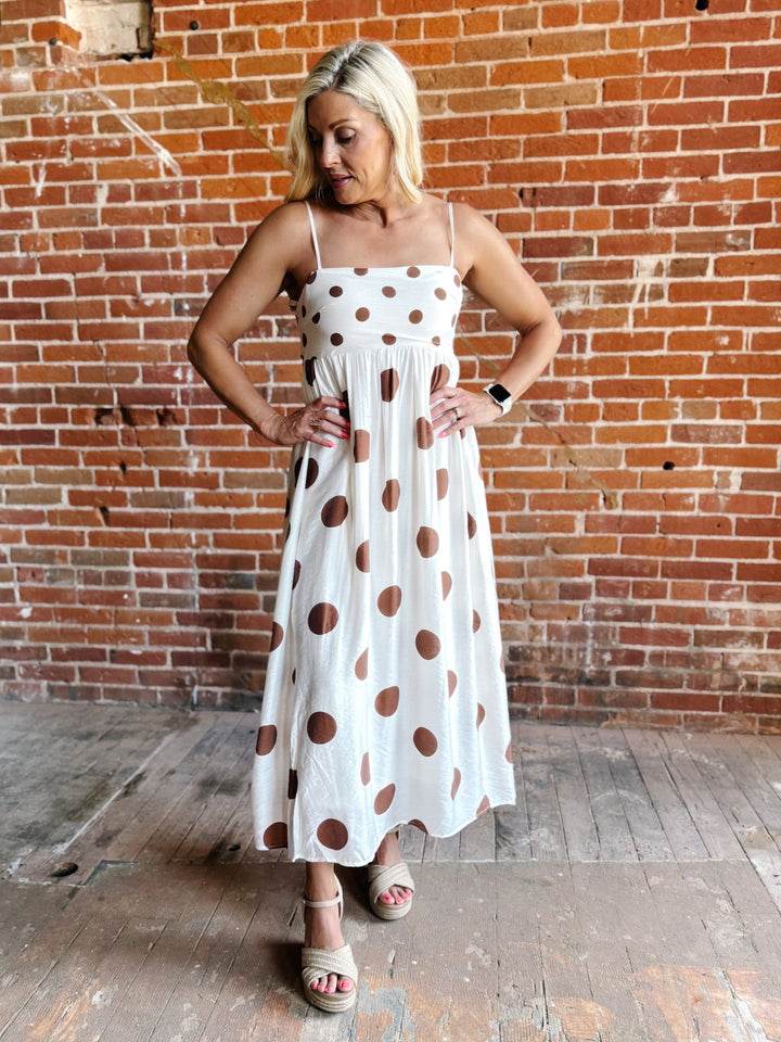 Woman wearing a white midi dress with oversized mocha polka dots, adjustable spaghetti straps, and an empire waist.