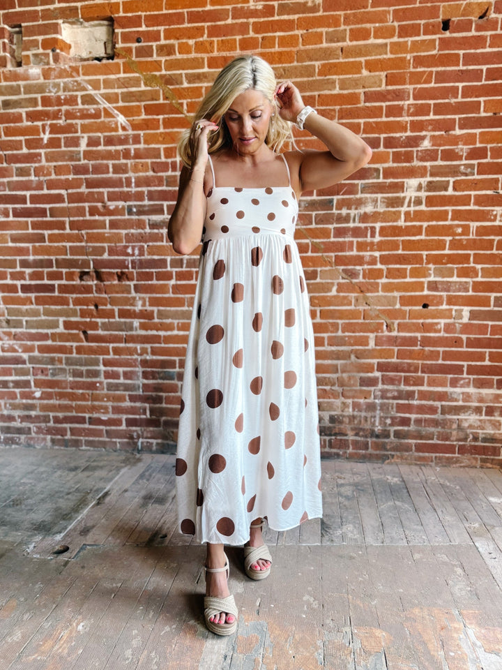 Woman wearing a white midi dress with oversized mocha polka dots, adjustable spaghetti straps, and an empire waist.