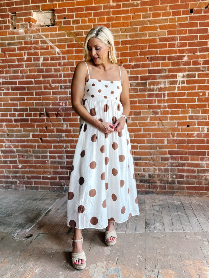 Woman wearing a white midi dress with oversized mocha polka dots, adjustable spaghetti straps, and an empire waist.