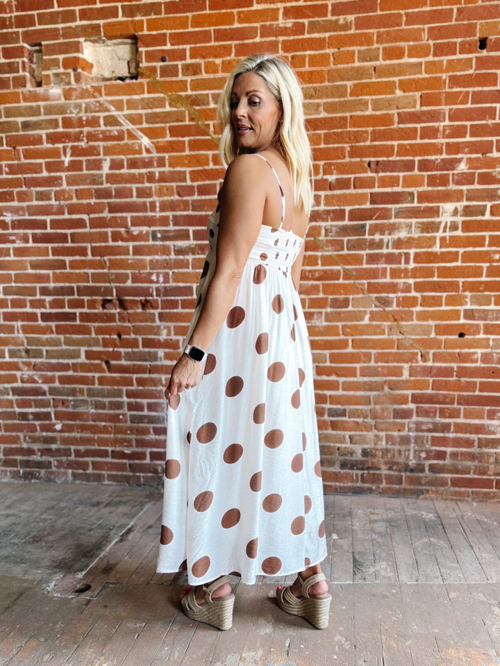 Woman wearing a white midi dress with oversized mocha polka dots, adjustable spaghetti straps, and an empire waist.