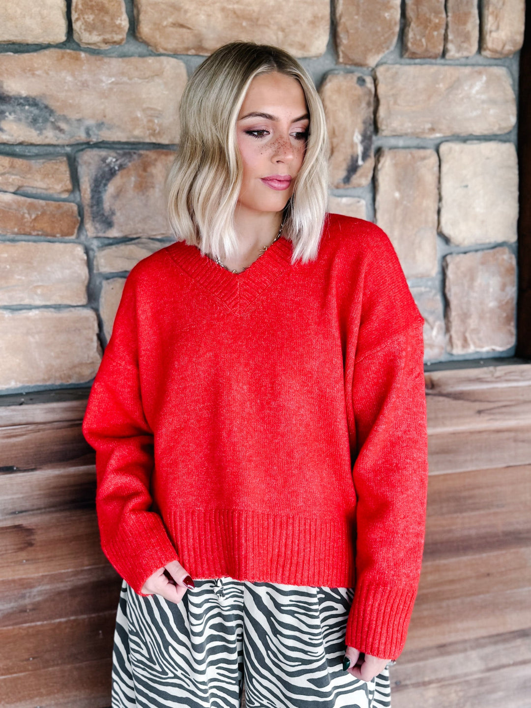 A woman with blonde wavy hair wearing a bright red knit sweater and brown-and-white zebra print pants, standing in front of a rustic stone wall.