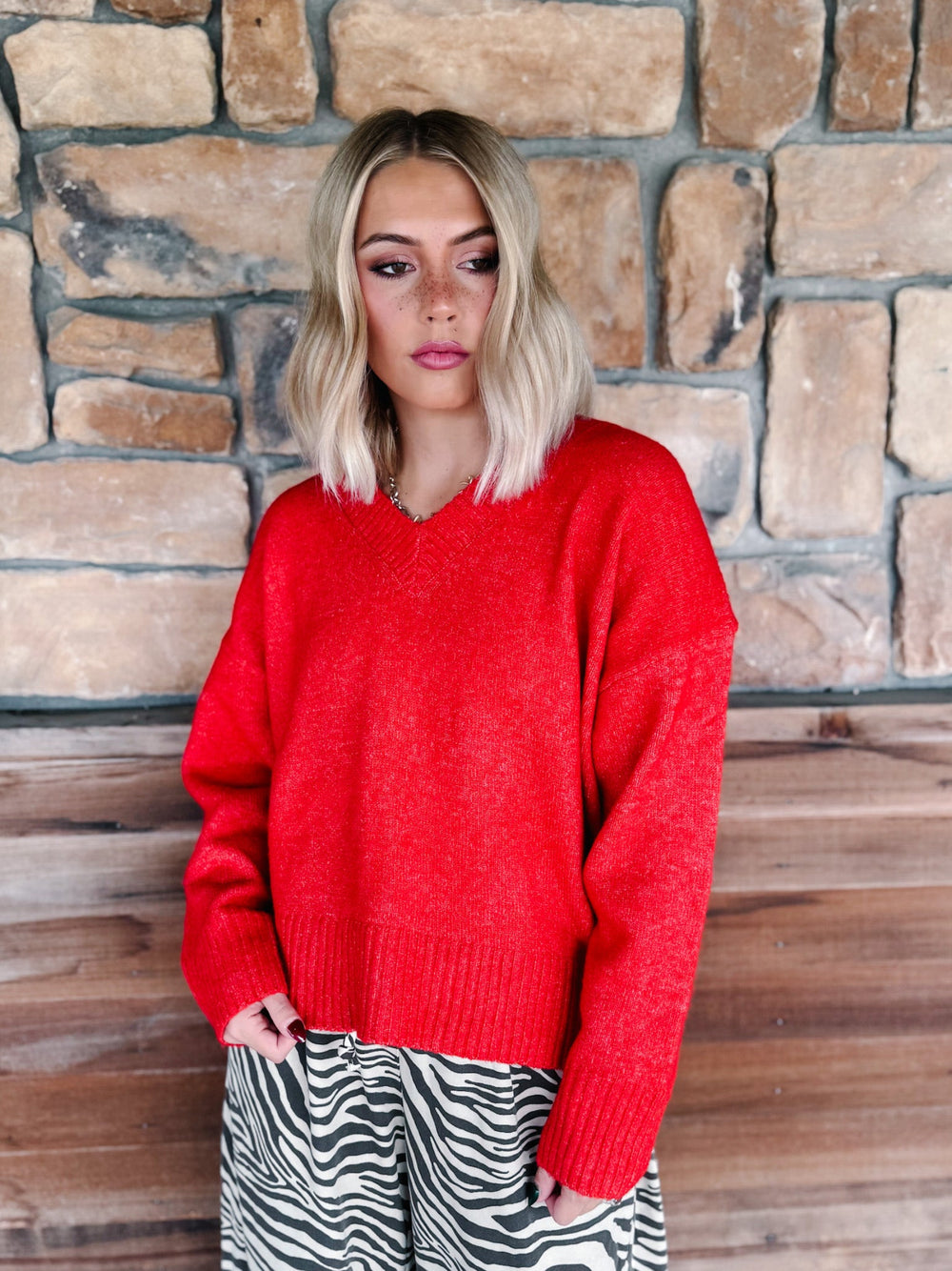 A woman with blonde wavy hair wearing a bright red knit sweater and brown-and-white zebra print pants, standing in front of a rustic stone wall.