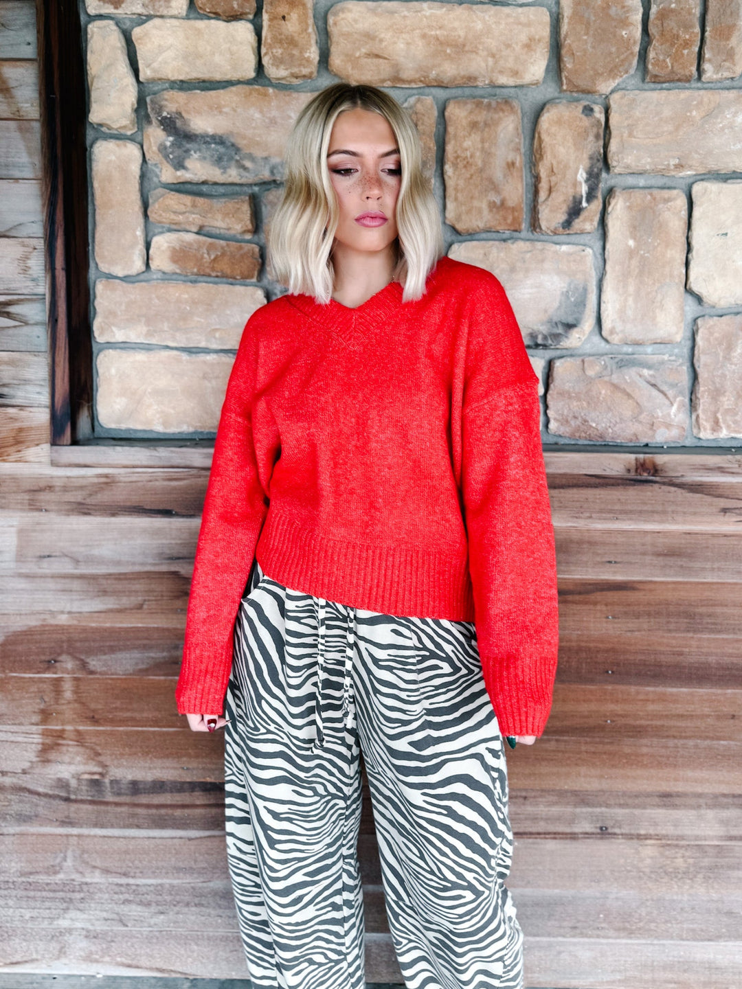 A woman with blonde wavy hair wearing a bright red knit sweater and brown-and-white zebra print pants, standing in front of a rustic stone wall.
