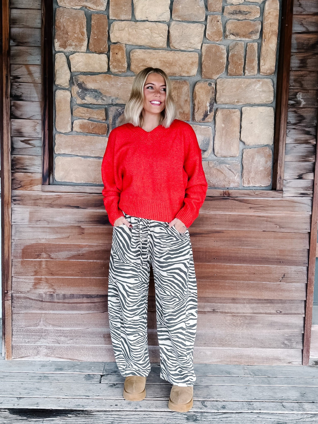 A woman with blonde wavy hair wearing a bright red knit sweater and brown-and-white zebra print pants, standing in front of a rustic stone wall.