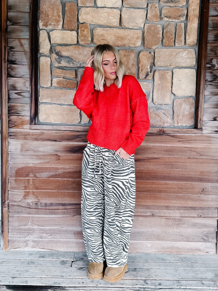 A woman with blonde wavy hair wearing a bright red knit sweater and brown-and-white zebra print pants, standing in front of a rustic stone wall.