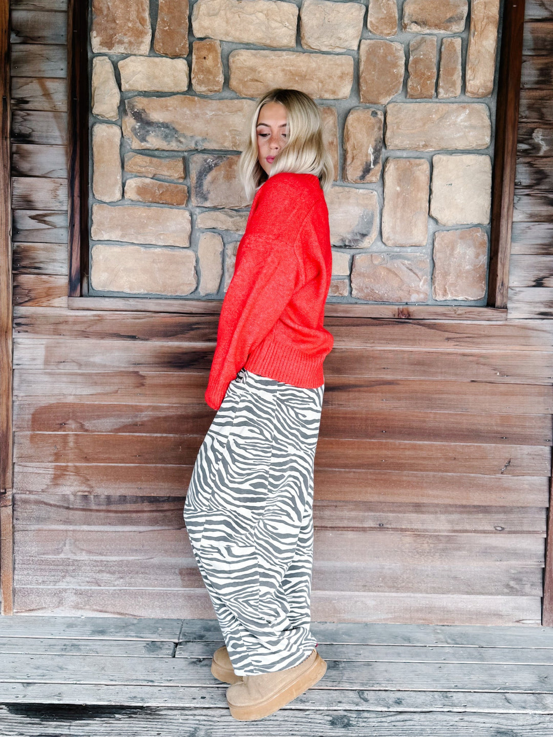 A woman with blonde wavy hair wearing a bright red knit sweater and brown-and-white zebra print pants, standing in front of a rustic stone wall.