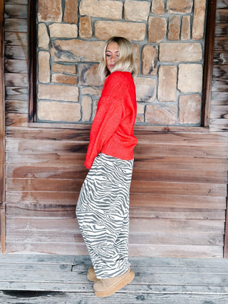 A woman with blonde wavy hair wearing a bright red knit sweater and brown-and-white zebra print pants, standing in front of a rustic stone wall.