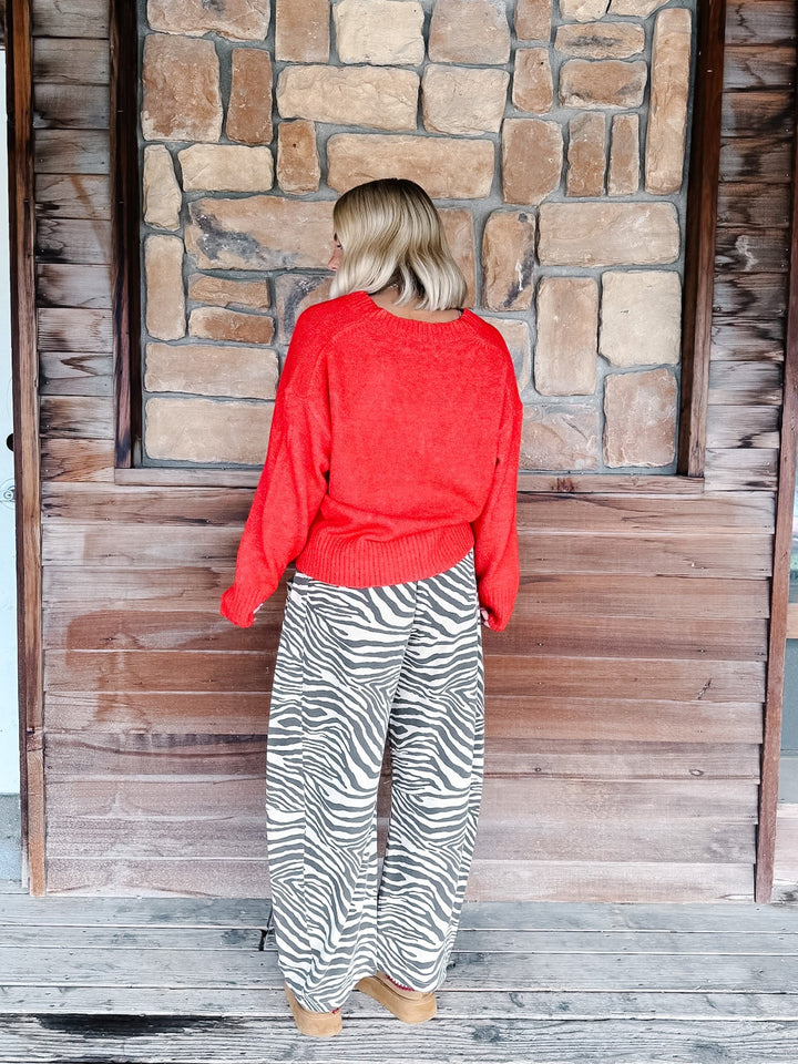 A woman with blonde wavy hair wearing a bright red knit sweater and brown-and-white zebra print pants, standing in front of a rustic stone wall.
