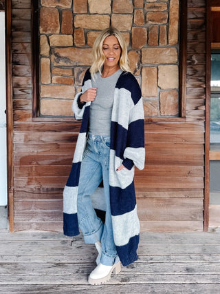 A woman stands on a wooden porch in front of a stone wall, wearing a long navy and light gray striped knit cardigan over a gray top, light blue jeans, and white boots.