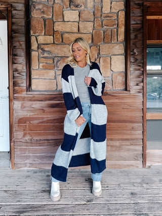 A woman stands on a wooden porch in front of a stone wall, wearing a long navy and light gray striped knit cardigan over a gray top, light blue jeans, and white boots.