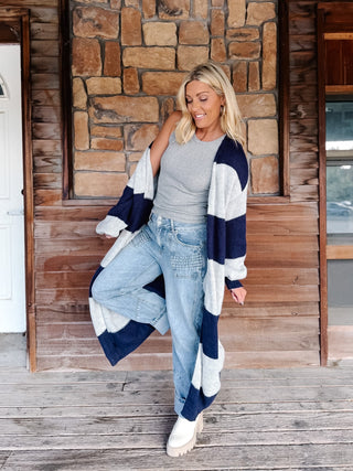 A woman stands on a wooden porch in front of a stone wall, wearing a long navy and light gray striped knit cardigan over a gray top, light blue jeans, and white boots.