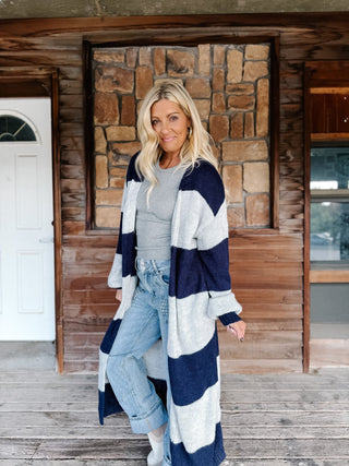 A woman stands on a wooden porch in front of a stone wall, wearing a long navy and light gray striped knit cardigan over a gray top, light blue jeans, and white boots.