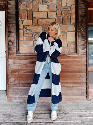 A woman stands on a wooden porch in front of a stone wall, wearing a long navy and light gray striped knit cardigan over a gray top, light blue jeans, and white boots.