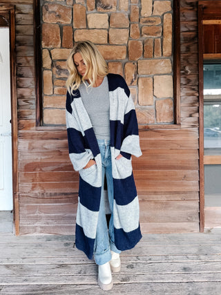 A woman stands on a wooden porch in front of a stone wall, wearing a long navy and light gray striped knit cardigan over a gray top, light blue jeans, and white boots.