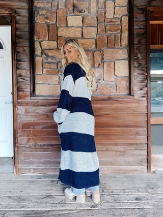 A woman stands on a wooden porch in front of a stone wall, wearing a long navy and light gray striped knit cardigan over a gray top, light blue jeans, and white boots.