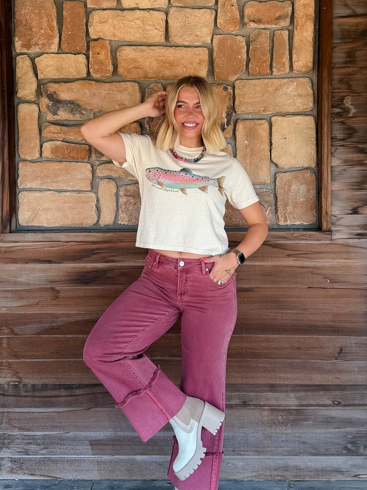 A woman posing in front of a rustic stone and wood wall, wearing a cream-colored graphic t-shirt with a fish design and dusty rose cropped jeans.