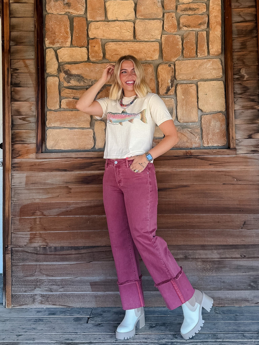 A woman posing in front of a rustic stone and wood wall, wearing a cream graphic t-shirt, dusty rose rolled-calf jeans, and white platform ankle boots.