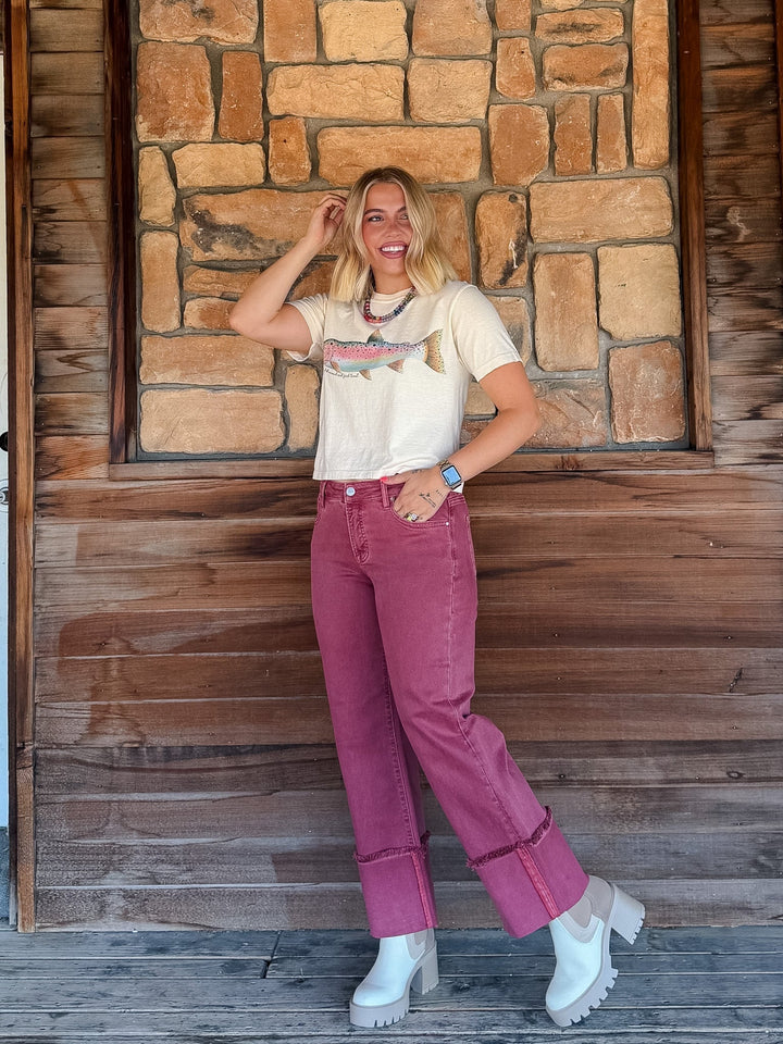 A woman posing in front of a rustic stone and wood wall, wearing a cream graphic t-shirt, dusty rose rolled-calf jeans, and white platform ankle boots.