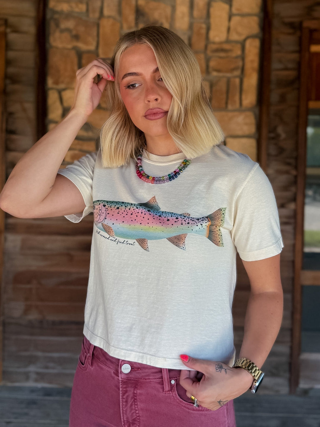 A woman with blonde hair wearing a cream-colored graphic t-shirt featuring a colorful rainbow trout, paired with dusty rose jeans, standing in front of a rustic stone wall.