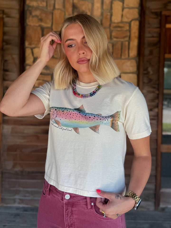 A woman with blonde hair wearing a cream-colored graphic t-shirt featuring a colorful rainbow trout, paired with dusty rose jeans, standing in front of a rustic stone wall.