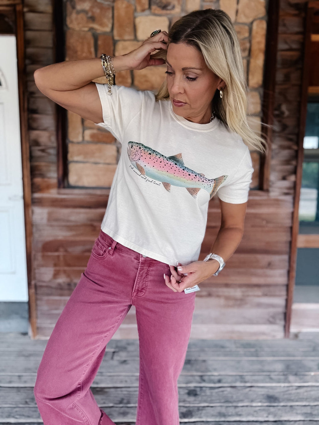 A woman wearing a white graphic t-shirt with a trout print and dusty rose jeans, standing on a wooden porch.