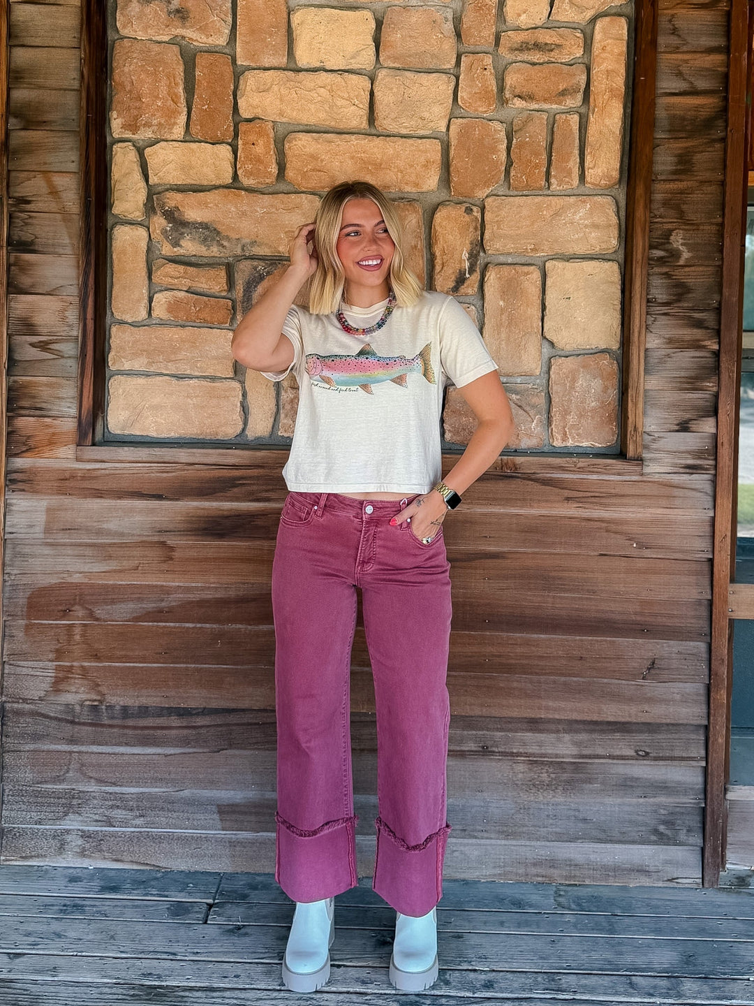 A woman stands on a wooden porch in front of a stone wall, wearing a cream graphic t-shirt with a fish design and dusty rose corduroy pants.