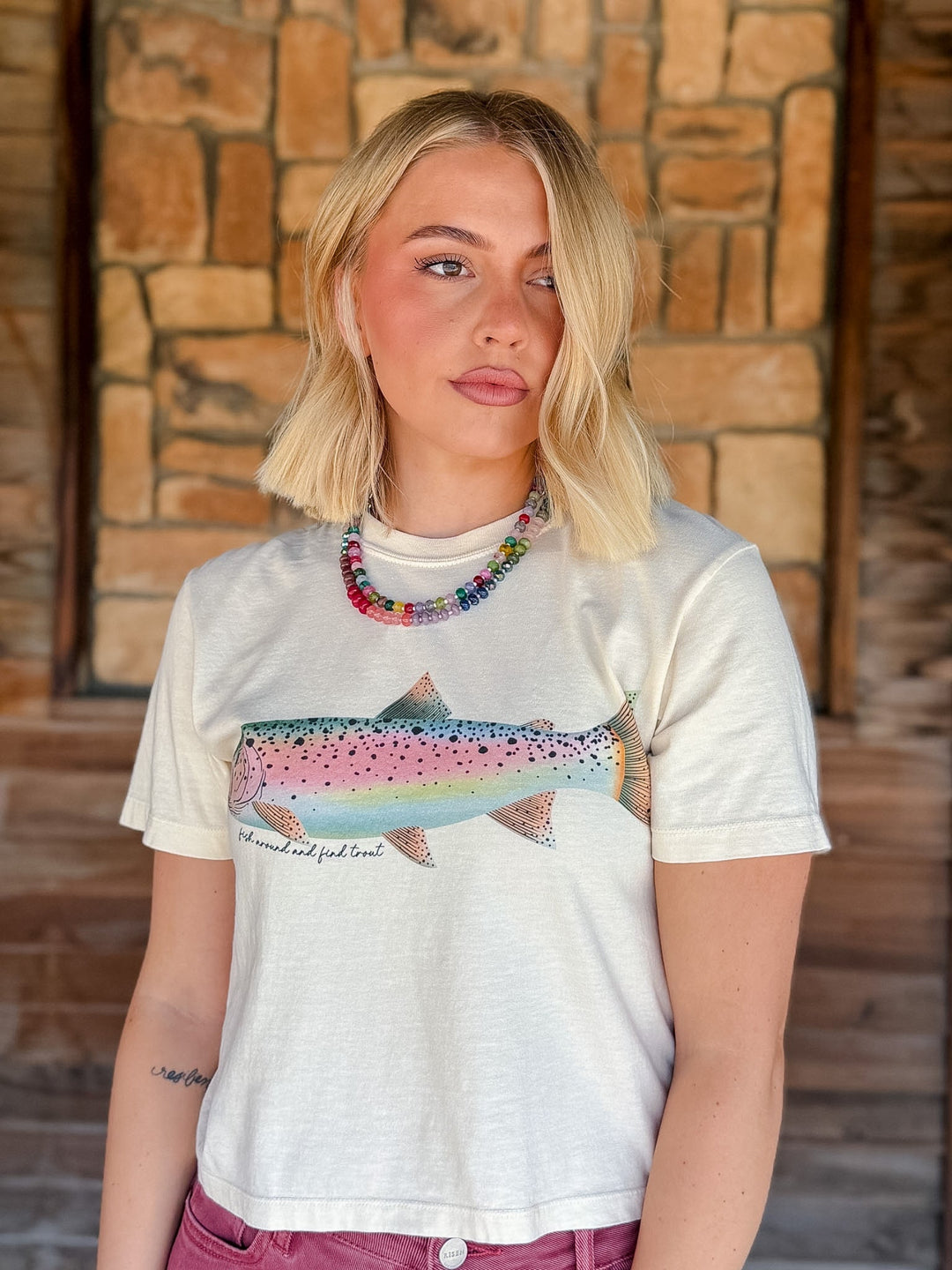 A blonde woman wearing a white graphic t-shirt with a colorful rainbow trout print and the text &
