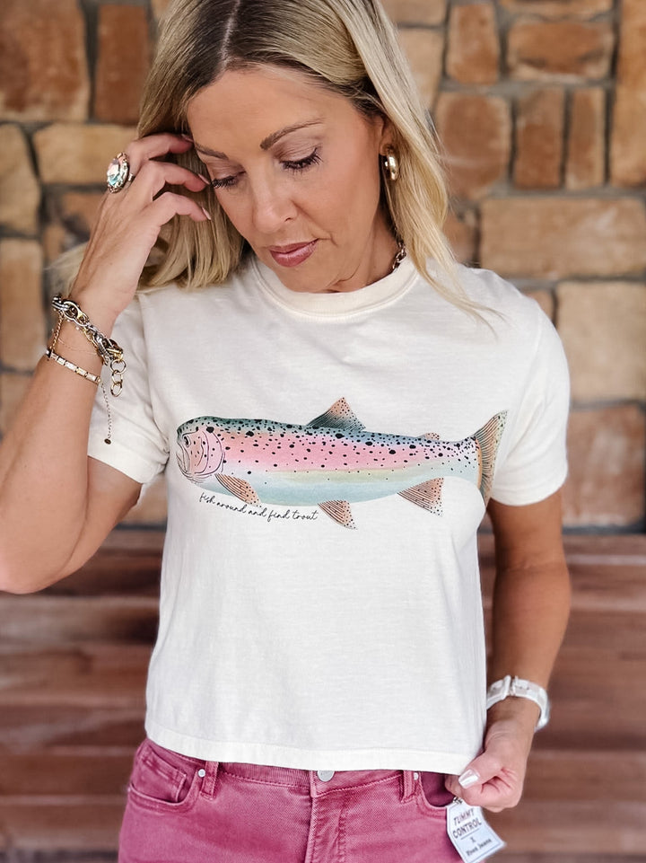 A woman wearing a white graphic t-shirt with a pink and blue trout design, paired with dusty rose jeans, standing in front of a rustic brick wall.
