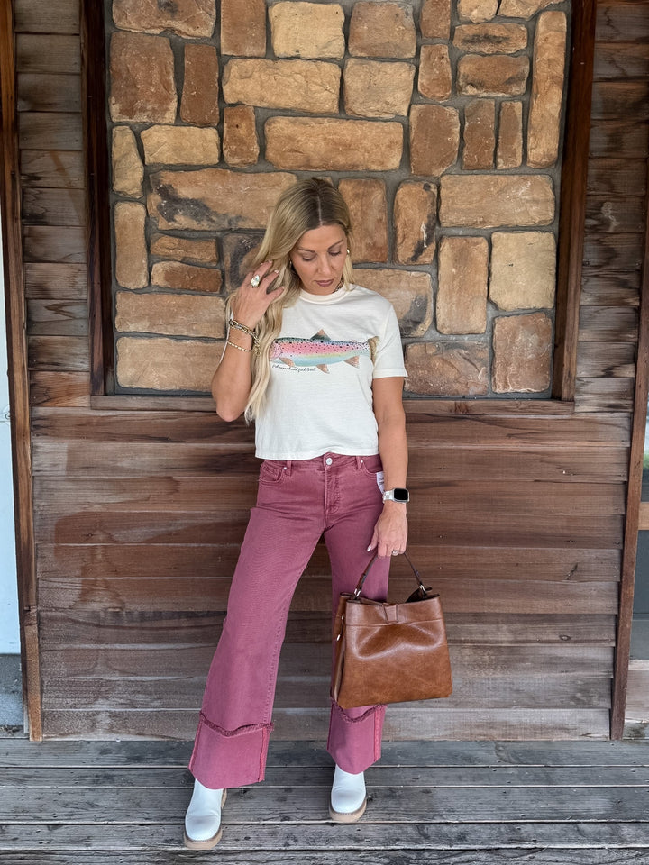 A woman stands in front of a rustic stone and wood wall, wearing a white graphic t-shirt, dusty rose flare jeans, white boots, and holding a brown leather handbag.