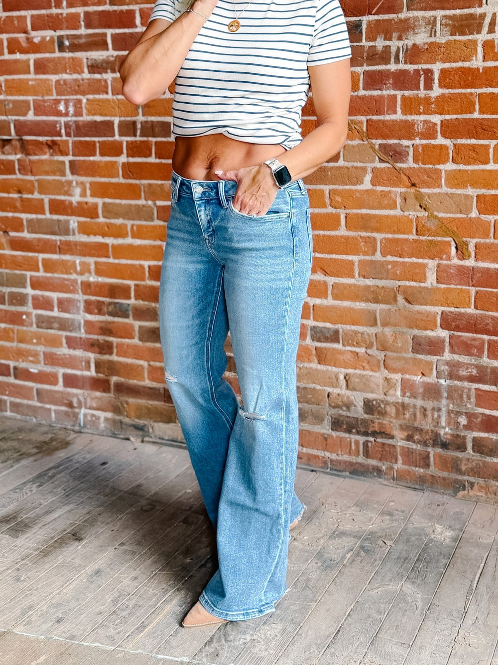 A person wearing light-wash blue flare jeans and a white cropped top with thin dark horizontal stripes, standing against a red-brown brick wall and wooden floor.