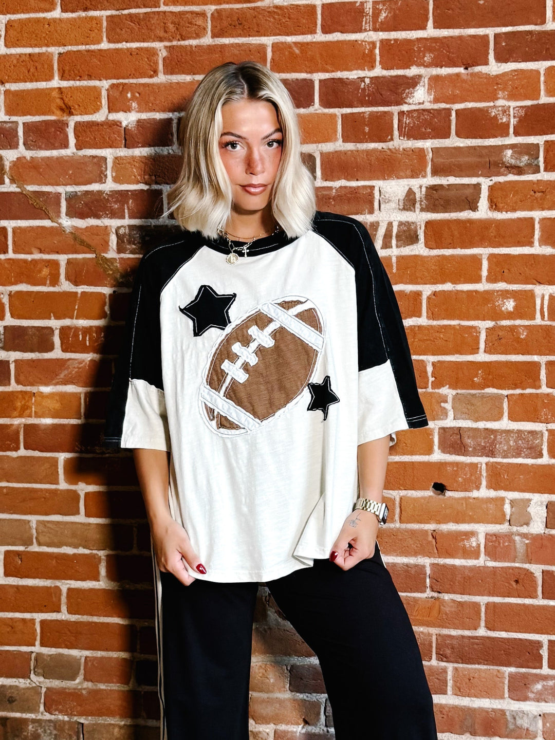 A woman with blonde hair stands against a red brick wall wearing a white and black raglan-style t-shirt with a brown football graphic and black star patches, paired with black pants.