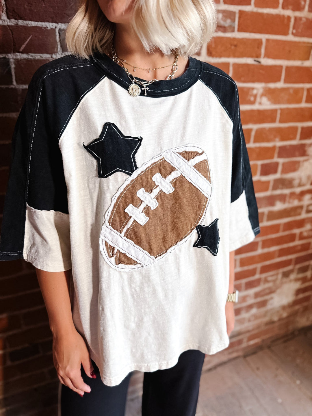 A person wearing a raglan-style short-sleeve T-shirt with a large applique football and two star motifs on the front. The shirt has black sleeves and shoulder yokes with an off-white body, shown against a brick wall background.