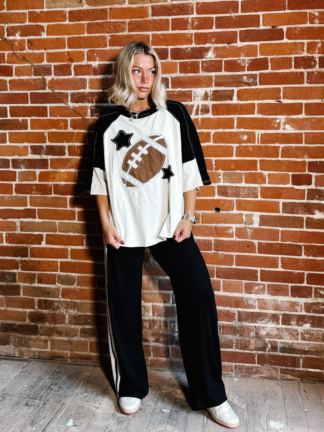 A woman stands against a red brick wall wearing a white and black raglan-style t-shirt with a football patch graphic, black wide-leg pants with white side stripes, and white sneakers.