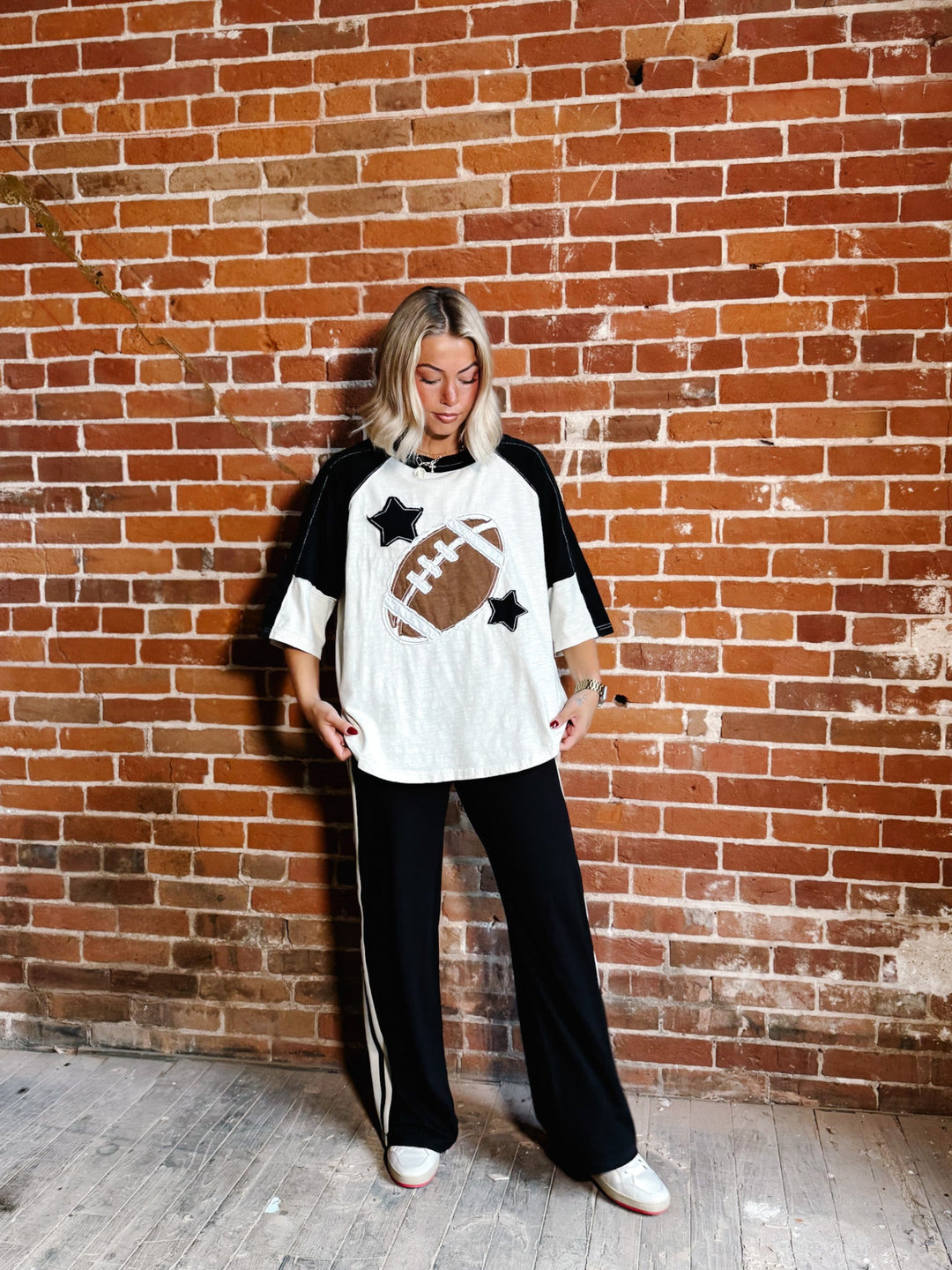 A woman stands against a red brick wall wearing a white and black raglan-style t-shirt with a brown football graphic and black stars, paired with black pants featuring white side stripes.