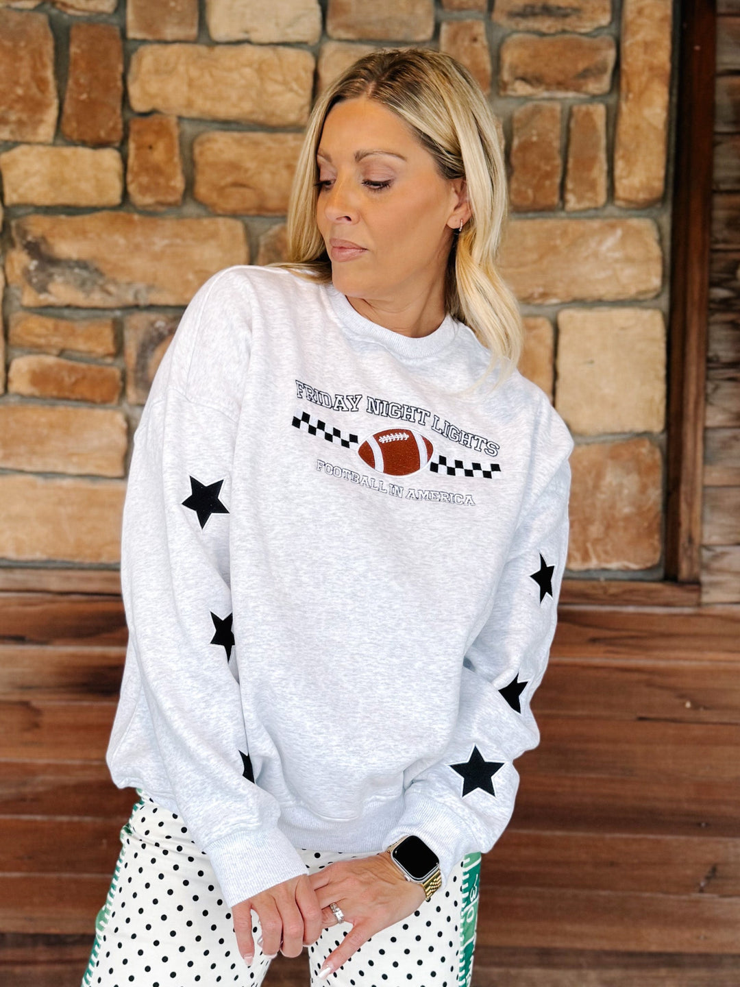 A woman wearing a light gray crewneck sweatshirt with black star prints and a football graphic that reads &