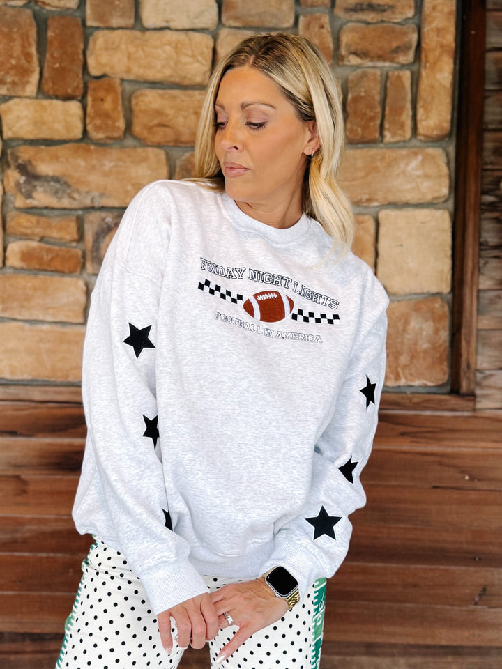 A woman wearing a light gray crewneck sweatshirt with black star prints and a football graphic that reads &