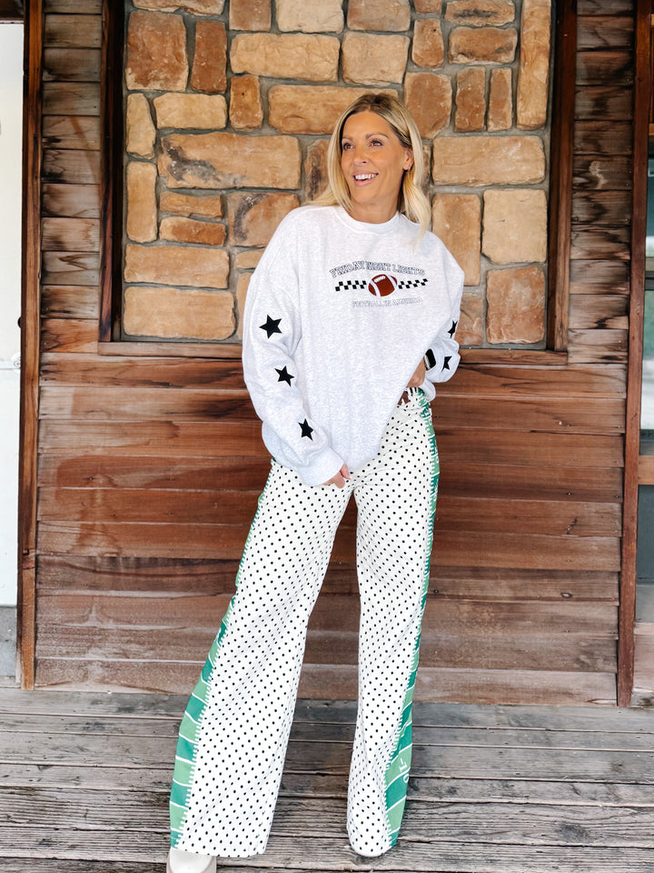 A woman stands on a wooden porch in front of a stone wall, wearing a white graphic sweatshirt with black stars and white wide-leg pants with black polka dots and green side accents.