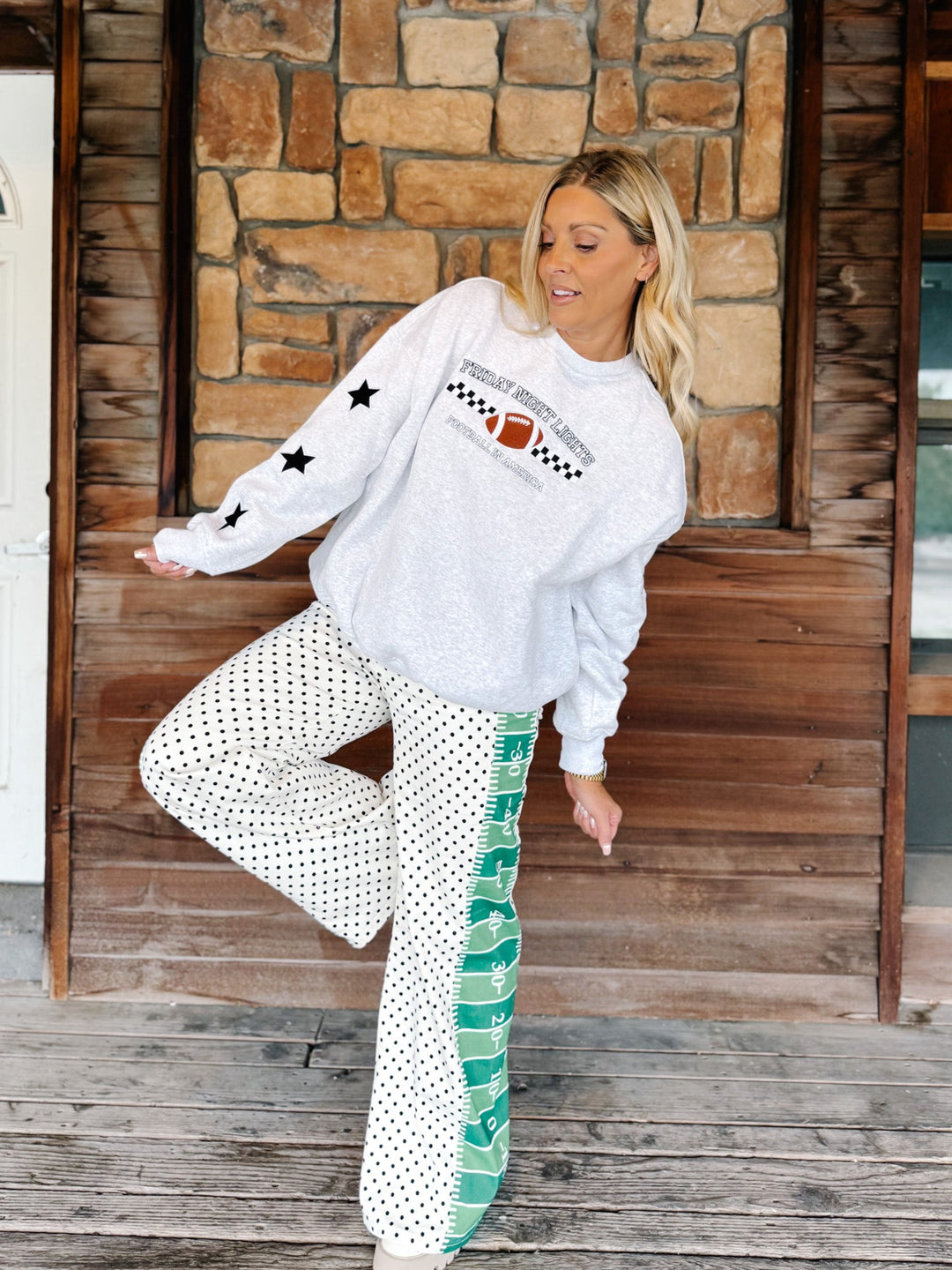A woman posing on a wooden porch wearing a light gray sweatshirt with a football graphic and black stars on the sleeve, paired with white pants featuring black polka dots and a green side stripe.
