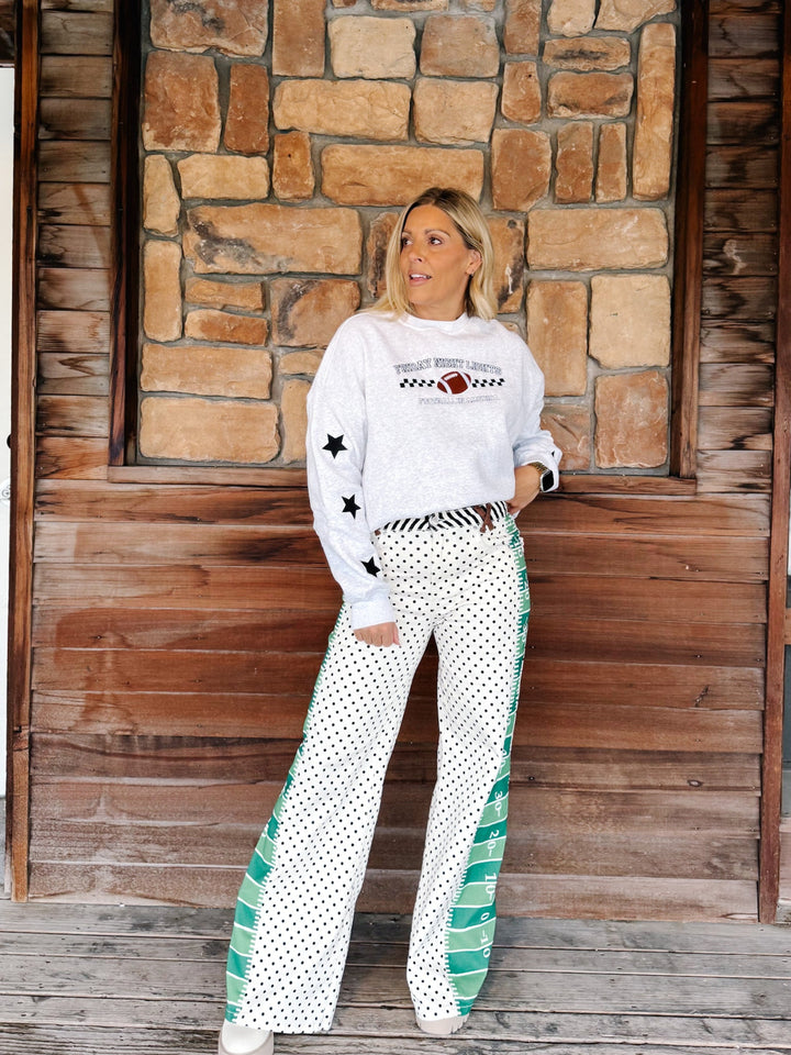 A woman stands on a wooden porch in front of a stone wall, wearing a white long-sleeve top with a football graphic and black stars, paired with white wide-leg pants featuring black polka dots and green side panels.