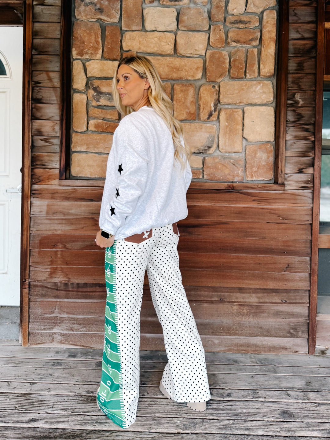A woman stands on a wooden porch in front of a stone and wood wall, wearing a white bomber-style jacket with black star details on the sleeves and white wide-leg pants with black polka dots and a green-and-white patterned side panel.