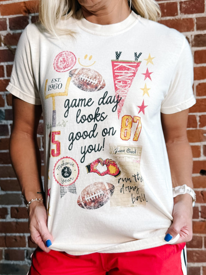 A woman wearing a cream-colored graphic t-shirt with football-themed designs and text, paired with red shorts, standing against a brick wall.