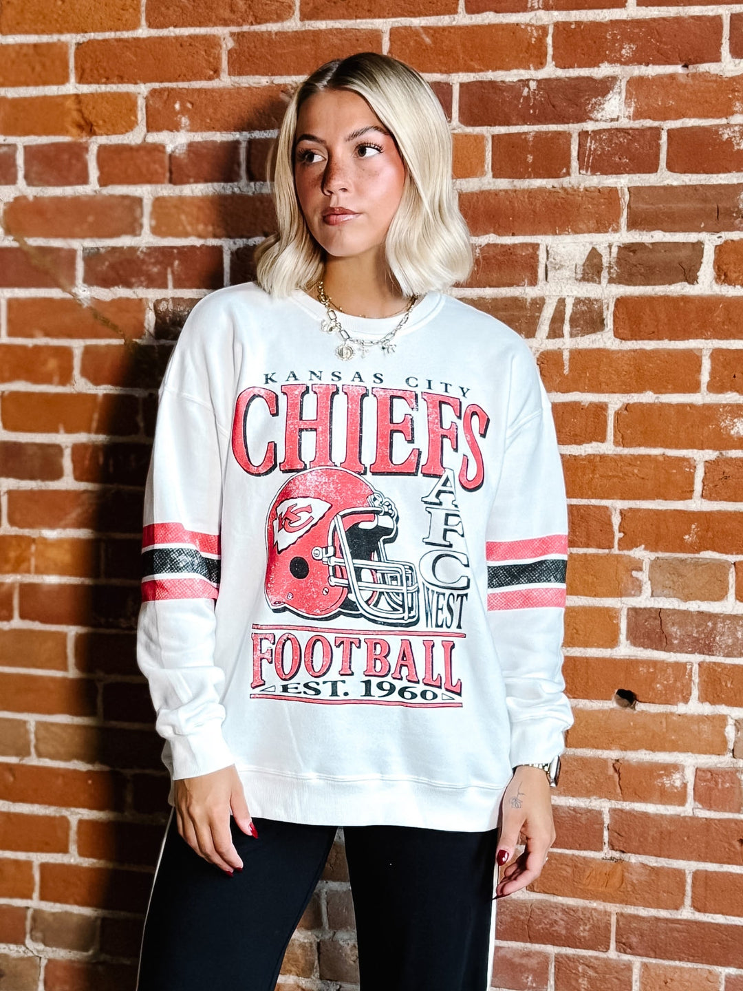 A woman with blonde hair stands against a red brick wall wearing a white Kansas City Chiefs football sweatshirt with red and black graphics.