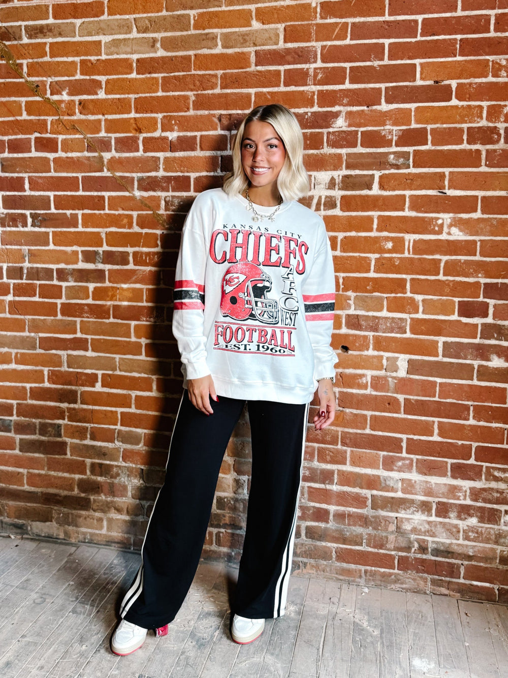 A woman stands against a red brick wall wearing a white long-sleeve sweatshirt with a red and black Chicago Chiefs football graphic, paired with black track pants featuring white side stripes and white sneakers.