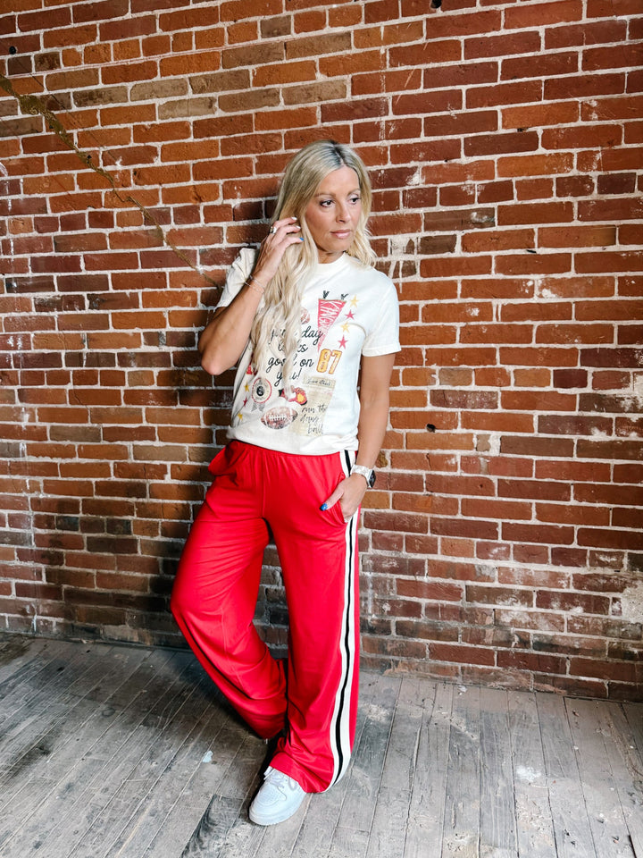 A woman posing in front of a red brick wall wearing a white graphic t-shirt and bright red track pants with white side stripes.